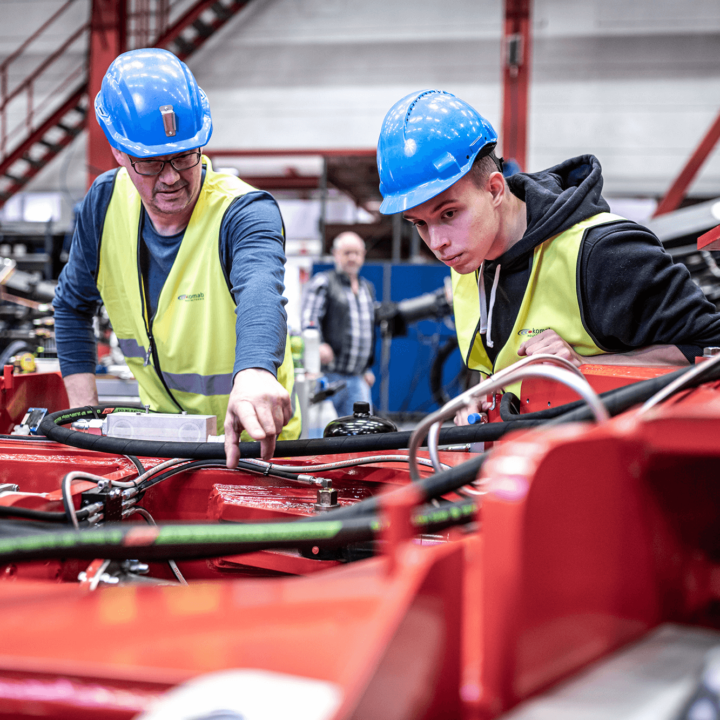 Two men with yellow vests and blue helmets in a factory looking at hoses in a red machine