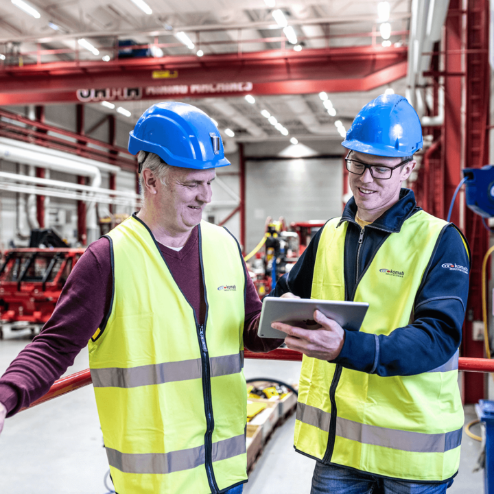 Two men with yellow vests and blue helmets in factory looking at tablet